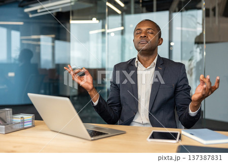 Mature man in a suit meditating at his office desk with eyes closed and hands in a mudra gesture, finding calm and focus amidst the contemporary workspace 137387831