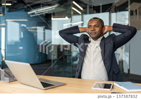 Mature businessman sitting at an office desk, stretching his arms behind his head, taking a moment to relax and clear his mind during a busy workday in a modern corporate environment Mature businessman sitting at an office desk, stretching his arms behind his head, taking a moment to relax and clear his mind during a busy workday in a modern corporate environment 137387832