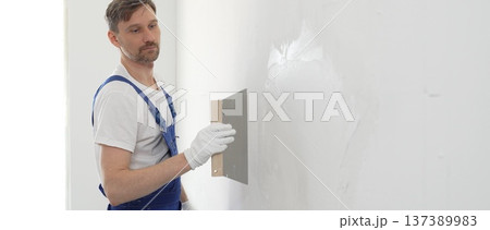 Male construction worker wearing protective gloves and blue construction coveralls, spreading plaster smoothly across wall using professional drywall taping knife during renovation project. Portrait Male construction worker wearing protective gloves and blue construction coveralls, spreading plaster smoothly across wall using professional drywall taping knife during renovation project. Portrait 137389983