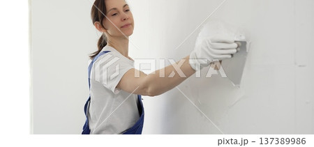 Female construction worker spreading finishing putty on white wall, wearing protective gloves and blue work overall while smoothing surface with putty knife for precise home repair 137389986