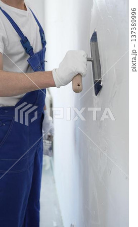 Professional construction worker wearing blue work overall is using a finishing trowel to carefully apply plaster on a wall, demonstrating expertise in home renovation 137389996