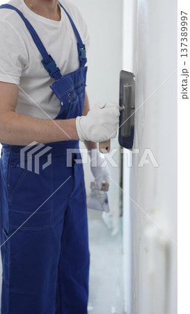 Professional construction worker wearing blue work overall is using a finishing trowel to carefully apply plaster on a wall, demonstrating expertise in home renovation 137389997