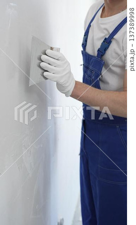 Close-up of man construction worker wearing gloves and blue overalls is using a putty knife to apply plaster on a white wall, performing home renovation and improvement Close-up of man construction worker wearing gloves and blue overalls is using a putty knife to apply plaster on a white wall, performing home renovation and improvement 137389998
