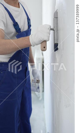 Professional construction worker wearing blue work overall is using a finishing trowel to carefully apply plaster on a wall, demonstrating expertise in home renovation Professional construction worker wearing blue work overall is using a finishing trowel to carefully apply plaster on a wall, demonstrating expertise in home renovation 137389999