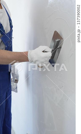 Professional construction worker wearing blue work overall is using a finishing trowel to carefully apply plaster on a wall, demonstrating expertise in home renovation Professional construction worker wearing blue work overall is using a finishing trowel to carefully apply plaster on a wall, demonstrating expertise in home renovation 137390002