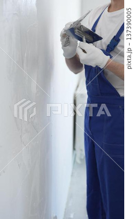 Professional construction worker wearing blue work overall is using a finishing trowel to carefully apply plaster on a wall, demonstrating expertise in home renovation Professional construction worker wearing blue work overall is using a finishing trowel to carefully apply plaster on a wall, demonstrating expertise in home renovation 137390005