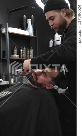 Professional barber carefully shaving client's beard with straight razor, applying gel and using hot towel in contemporary barbershop setting, vertical view Professional barber carefully shaving client's beard with straight razor, applying gel and using hot towel in contemporary barbershop setting, vertical view 137390024