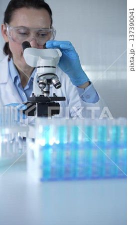 Female scientist wearing lab coat and protective blue gloves using microscope doing research in modern laboratory, portrait view. Low key lighting photo. Science and Medicine Female scientist wearing lab coat and protective blue gloves using microscope doing research in modern laboratory, portrait view. Low key lighting photo. Science and Medicine 137390041