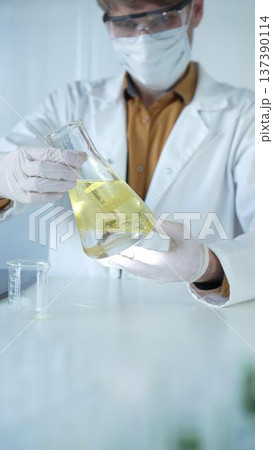 Male scientist wearing white protective gloves, mask and glasses is holding a yellow chemical solution inside an Erlenmeyer flask in a laboratory, vertical portrait. Science and medicine Male scientist wearing white protective gloves, mask and glasses is holding a yellow chemical solution inside an Erlenmeyer flask in a laboratory, vertical portrait. Science and medicine 137390114
