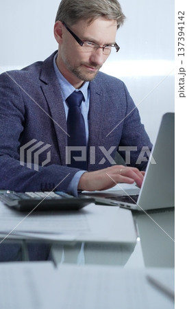 Businessman wearing blue suit is working with laptop, calculator and documents at the glass table in office set. Low lighting, vertical portrait. Business people and audit Businessman wearing blue suit is working with laptop, calculator and documents at the glass table in office set. Low lighting, vertical portrait. Business people and audit 137394142