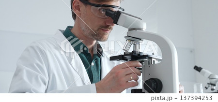 Adult male scientist wearing lab coat and protective glasses using microscope, doing scientific research in modern laboratory. Medicine, healthcare and science concept Adult male scientist wearing lab coat and protective glasses using microscope, doing scientific research in modern laboratory. Medicine, healthcare and science concept 137394253