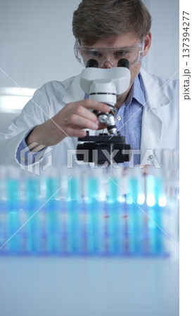Male scientist researcher wearing lab coat and protective glasses, is using microscope in modern laboratory setup, portrait view. Low key lighting photo. Science concept 137394277