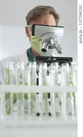 Male scientific researcher in green protective gear analyzing specimens through a microscope in a sterile laboratory environment. Medicine and science concept 137394282