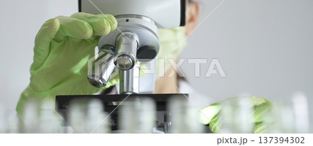 Close up of scientific precision involving green gloved hands adjusting microscope lens, blurred laboratory glassware highlighting medical investigation. Medicine, laboratory and science concept Close up of scientific precision involving green gloved hands adjusting microscope lens, blurred laboratory glassware highlighting medical investigation. Medicine, laboratory and science concept 137394302