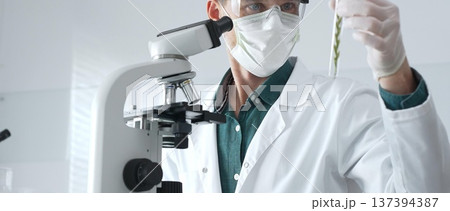 Masked adult man researcher examining green botanical samples in test tube in a sterile laboratory during biological analysis. Medicine, healthcare and science concept 137394387