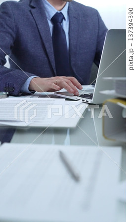 Businessman wearing blue suit is working with laptop, calculator and documents at the glass table in office set. Low lighting, close-up vertical view. Business people and audit Businessman wearing blue suit is working with laptop, calculator and documents at the glass table in office set. Low lighting, close-up vertical view. Business people and audit 137394390