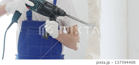 Close up of unrecognizable male construction worker wearing blue overalls and protective white gloves, is demolishing white wall with rotary hammer drill, generating dust, horizontal view. Renovation 137394400