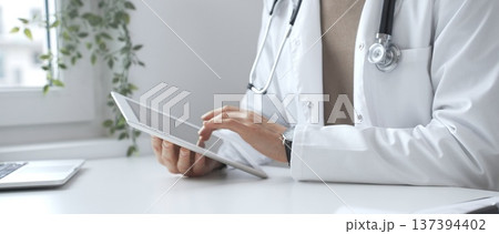 Doctor man wearing lab coat and stethoscope using digital tablet in modern medical office, browsing patient information or conducting online consultation. Medicine and health care 137394402