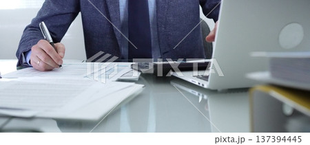 Businessman wearing blue suit is working with laptop, calculator and documents in a professional office setting. Low lighting, close-up view. Business people, audit concept Businessman wearing blue suit is working with laptop, calculator and documents in a professional office setting. Low lighting, close-up view. Business people, audit concept 137394445