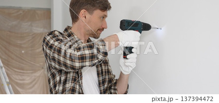 Middle aged man wearing checked shirt and white gloves, is drilling hole in a wall with cordless drill during home renovation work. Portrait view Middle aged man wearing checked shirt and white gloves, is drilling hole in a wall with cordless drill during home renovation work. Portrait view 137394472