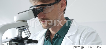 Adult male scientist wearing protective glasses and lab coat analyzing samples with a microscope in a modern laboratory setting. Medicine, healthcare and science concept 137394520