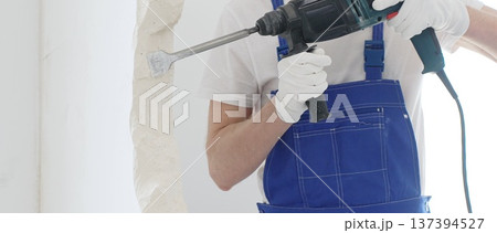 Unrecognizable male construction worker wearing blue overalls and protective white gloves, is demolishing white wall with rotary hammer drill, generating dust, close up horizontal view. Renovation Unrecognizable male construction worker wearing blue overalls and protective white gloves, is demolishing white wall with rotary hammer drill, generating dust, close up horizontal view. Renovation 137394527