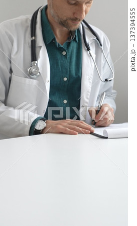 Doctor man wearing lab coat over green shirt, and stethoscope is writing medical prescription or filling medical documents sitting at office desk. Medicine and health care Doctor man wearing lab coat over green shirt, and stethoscope is writing medical prescription or filling medical documents sitting at office desk. Medicine and health care 137394555