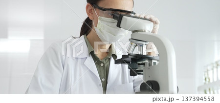 Female scientist in a lab coat and protective glasses examining samples through a microscope in a research laboratory. Medicine, healthcare and science concept 137394558