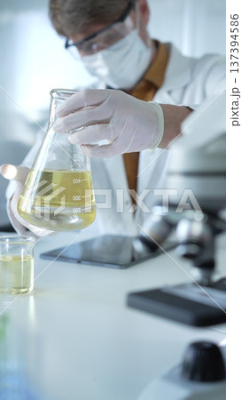 Man scientist researcher, wearing white protective gloves, mask and glasses, is holding a yellow chemical solution inside Erlenmeyer flask in a laboratory, vertical portrait view. Science and medicine Man scientist researcher, wearing white protective gloves, mask and glasses, is holding a yellow chemical solution inside Erlenmeyer flask in a laboratory, vertical portrait view. Science and medicine 137394586