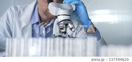 Male scientist researcher wearing blue gloves, protective glasses is using microscope in modern laboratory, portrait view. Low key lighting photo. Science and Medicine concepts Male scientist researcher wearing blue gloves, protective glasses is using microscope in modern laboratory, portrait view. Low key lighting photo. Science and Medicine concepts 137394620