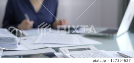 White calculator, pen and documents are lying on accountant's desk. Businesswoman wearing blue dress is working with documents on the background. Business and audit concept White calculator, pen and documents are lying on accountant's desk. Businesswoman wearing blue dress is working with documents on the background. Business and audit concept 137394627