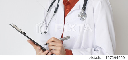 Close up of female doctor wearing stethoscope and white coat over brick red blouse is standing and writing on a clipboard, representing medical expertise and patient care. Medicine concept 137394660