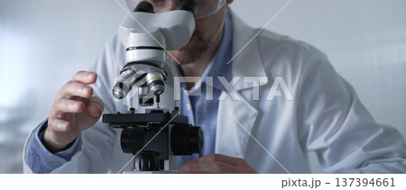 Male scientist wearing lab coat and protective glasses using microscope doing research in modern laboratory setup, portrait view. Low key lighting photo. Science and Medicine concept 137394661