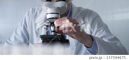 Male scientist wearing lab coat and protective glasses using microscope doing research in modern laboratory setup, portrait view. Low key lighting photo. Science and Medicine concept 137394675