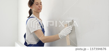 Woman construction worker wearing protective gloves and blue construction coveralls, spreading plaster smoothly across wall using professional drywall taping knife during renovation project. Portrait Woman construction worker wearing protective gloves and blue construction coveralls, spreading plaster smoothly across wall using professional drywall taping knife during renovation project. Portrait 137394676