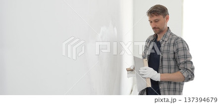 Male construction worker wearing protective gloves and plaid shirt, spreading plaster smoothly across wall surface using professional drywall taping knife during renovation project. Portrait view 137394679