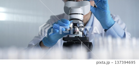 Woman scientist wearing lab coat, white protective gloves and glasses, is using microscope in modern laboratory setup, portrait view. Low key lighting photo. Science and Medicine Woman scientist wearing lab coat, white protective gloves and glasses, is using microscope in modern laboratory setup, portrait view. Low key lighting photo. Science and Medicine 137394695