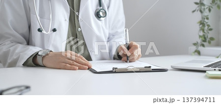 Professional doctor woman wearing white lab coat and stethoscope documenting patient information on clipboard near laptop in clinic workspace. Medicine and health care Professional doctor woman wearing white lab coat and stethoscope documenting patient information on clipboard near laptop in clinic workspace. Medicine and health care 137394711