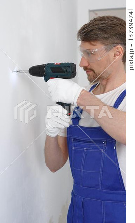 Professional male construction worker in a blue jumpsuit, safety goggles, and work gloves is screwing a screw into the wall using a cordless screwdriver, carrying out a careful home renovation Professional male construction worker in a blue jumpsuit, safety goggles, and work gloves is screwing a screw into the wall using a cordless screwdriver, carrying out a careful home renovation 137394741