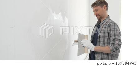 Man construction worker wearing protective gloves and plaid shirt, spreading plaster smoothly across wall surface using professional drywall taping knife during renovation project Man construction worker wearing protective gloves and plaid shirt, spreading plaster smoothly across wall surface using professional drywall taping knife during renovation project 137394743