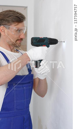 Professional male construction worker in a blue jumpsuit, safety goggles, and work gloves is screwing a screw into the wall using a cordless screwdriver. Home renovation concept Professional male construction worker in a blue jumpsuit, safety goggles, and work gloves is screwing a screw into the wall using a cordless screwdriver. Home renovation concept 137394757