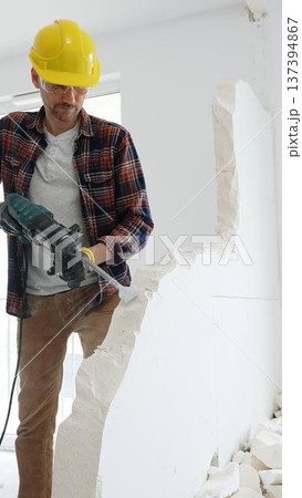 Male construction worker wearing red checkered shirt, yellow hard hat and protective gloves, is demolishing white wall with rotary hammer drill, generating dust, medium long shot. Renovation project Male construction worker wearing red checkered shirt, yellow hard hat and protective gloves, is demolishing white wall with rotary hammer drill, generating dust, medium long shot. Renovation project 137394867