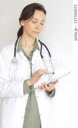Portrait of a doctor woman wearing white medical coat over green blouse, stethoscope, is reviewing patient records on digital tablet, standing in medical workspace with natural lighting. Medicine 137394872