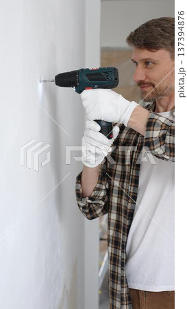 Middle aged man wearing checked shirt and white gloves, is drilling hole in a wall with cordless drill during home renovation work. Portrait view 137394876