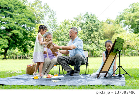 Asian senior man and woman express glad and look at the drawing of one girl while the other girl concentrate to draw or paint in public park with day light. Asian senior man and woman express glad and look at the drawing of one girl while the other girl concentrate to draw or paint in public park with day light. 137395480