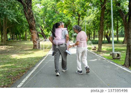 Back of Asian girl piggyback ride to senior man and walk together with senior woman along the road in public park and they look happy to relax with nature together. Back of Asian girl piggyback ride to senior man and walk together with senior woman along the road in public park and they look happy to relax with nature together. 137395496