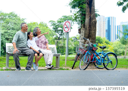 Asian grandfather and grandmother or senior man and woman look at the direction that the girl point to when they sit on garden bench in the public park with happiness. Asian grandfather and grandmother or senior man and woman look at the direction that the girl point to when they sit on garden bench in the public park with happiness. 137395519