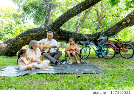 Front view of Asian grandfather and grandmother enjoy and relax with two girls in public park with eating fruit and play music and sing a song together. Front view of Asian grandfather and grandmother enjoy and relax with two girls in public park with eating fruit and play music and sing a song together. 137395521