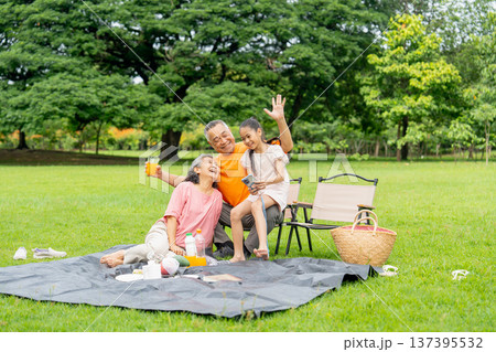 Asian little girl sit and show the picture that use mobile phone to take a photo with senior man and woman during relax with family in public park. Asian little girl sit and show the picture that use mobile phone to take a photo with senior man and woman during relax with family in public park. 137395532