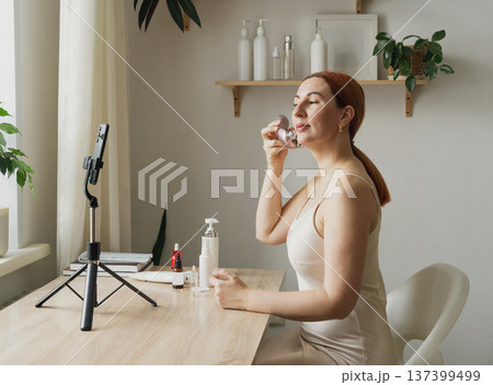 Woman creating beauty vlog applying facial massager. A beauty blogger records content for social media using a smartphone Woman creating beauty vlog applying facial massager. A beauty blogger records content for social media using a smartphone 137399499